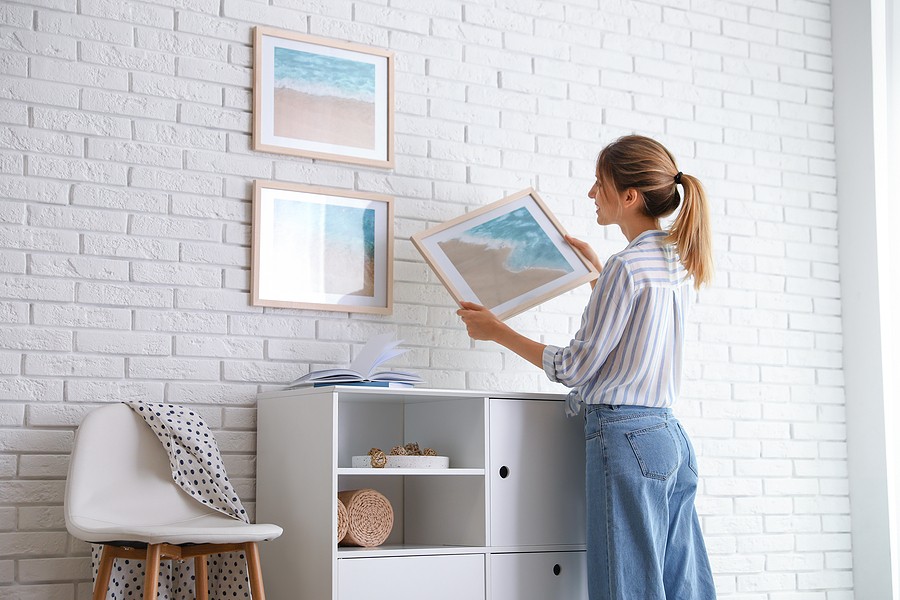 Woman hanging framed beach art on a white brick wall in a staged room.